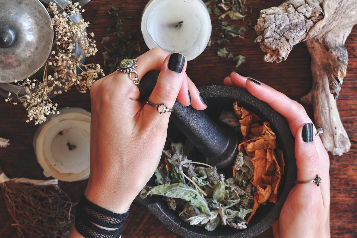 Kitchen witchery – female wiccan witch holding pestle and mortar in her hands, making magickal herb blend for a spell. Mix of dried herbs ready to be blended. Messy witch’s altar on dark wooden table Wiedźma, znachorka szykująca napar z ziół